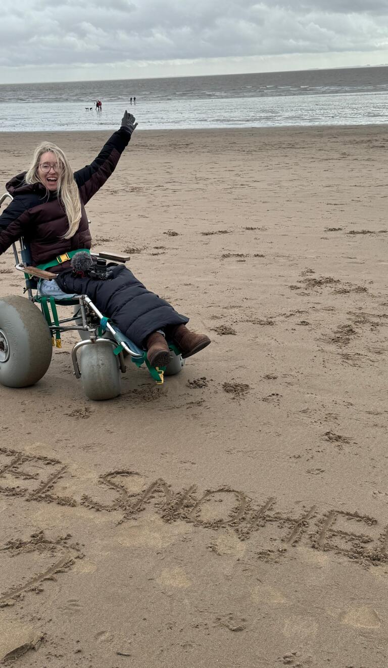 woman in beach wheelchair on sandy beach with words @WheelsnoHeels written in sand.