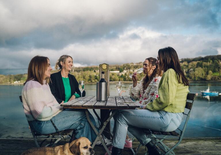 group of women drinking at outside table with dog at their feet and views of Menai.