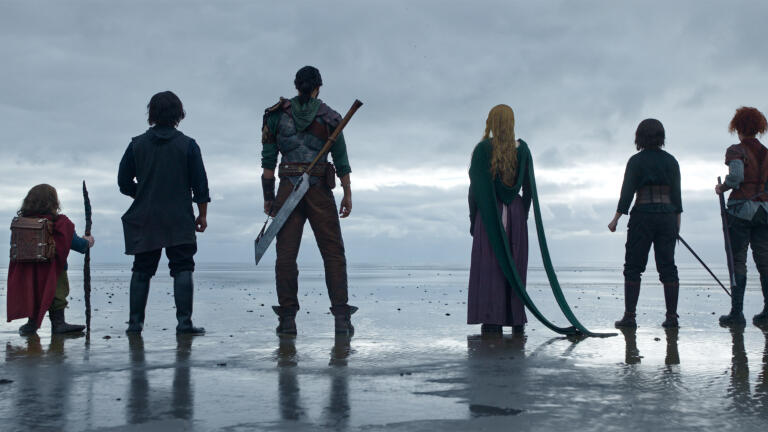 (L-R): Willow Ufgood (Warwick Davis), Graydon (Tony Revolori), Boorman (Amar Chadha-Patel), Dove (Ellie Bamber), Kit (Ruby Cruz) and Jade (Erin Kellyman) stood by the shoreline looking out across a grey sea and sky in Lucasfilm's Willow.