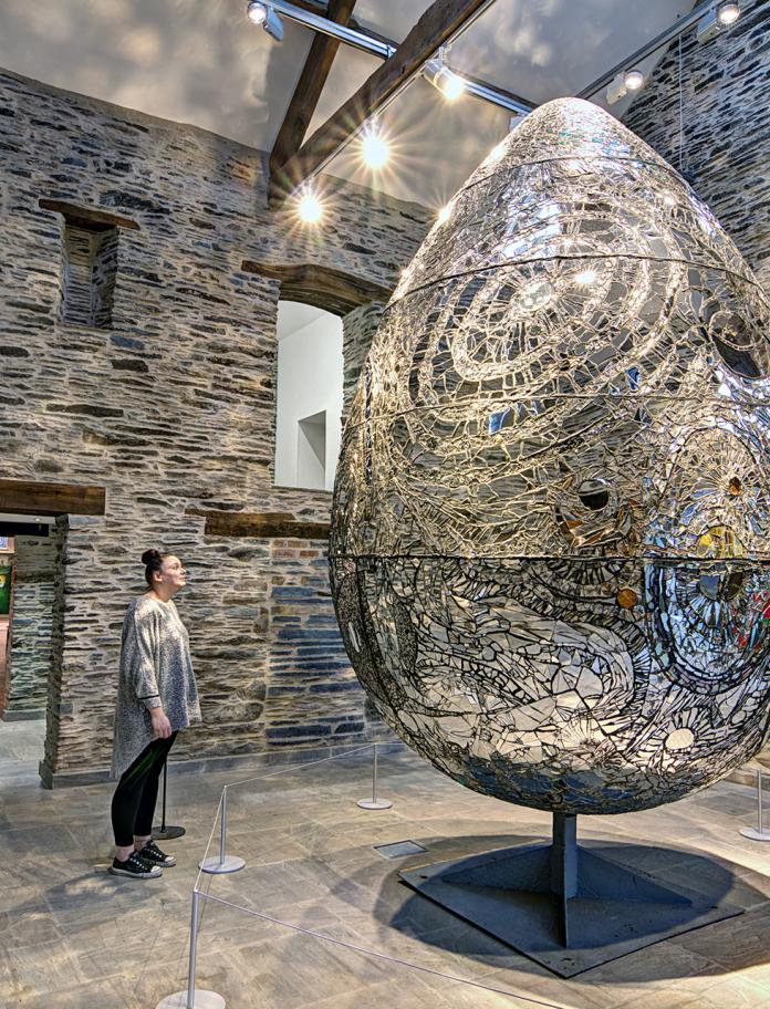 Giant egg sculpture with woman stood looking at it inside a stone wall room.