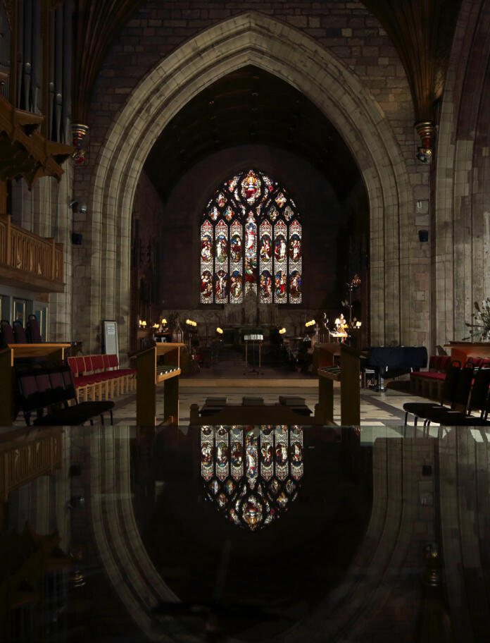 Das Innere einer Kathedrale mit steinernem Bogen und Bleiglasfenster, das sich auf dem Boden spiegelt.