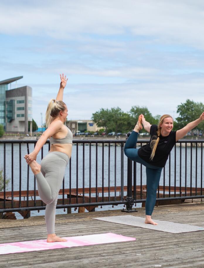 Two people performing stretching yoga poses on a waterside boardwalk with buildings in the background.