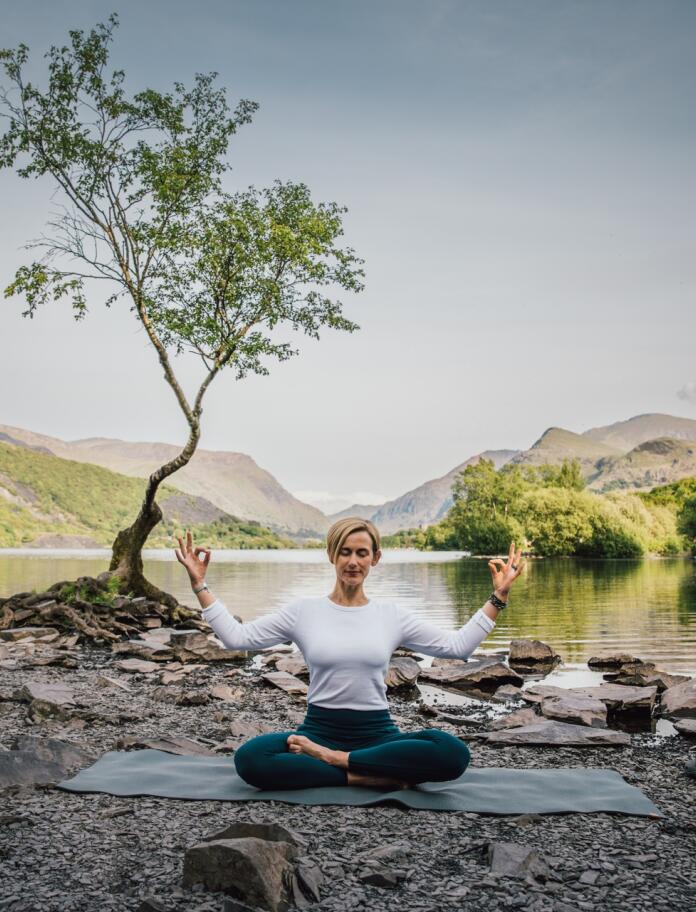 Person sat in a yoga pose in front of a lake with a tree to their left in the foreground and mountains in the distance.