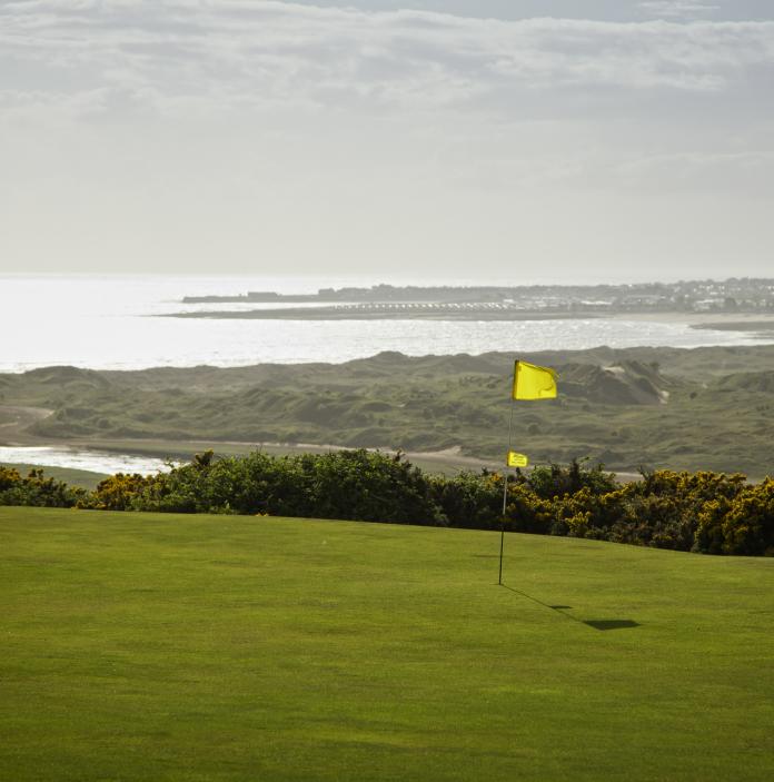 Ein Flaggenstock auf dem Grün des Southerndown Golf Clubs mit dem Meer und Landstrichen im Hintergrund.