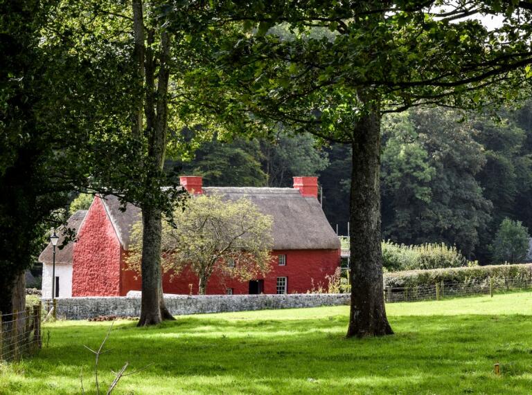 A red painted thatched cottage surrounded by trees and greenery.