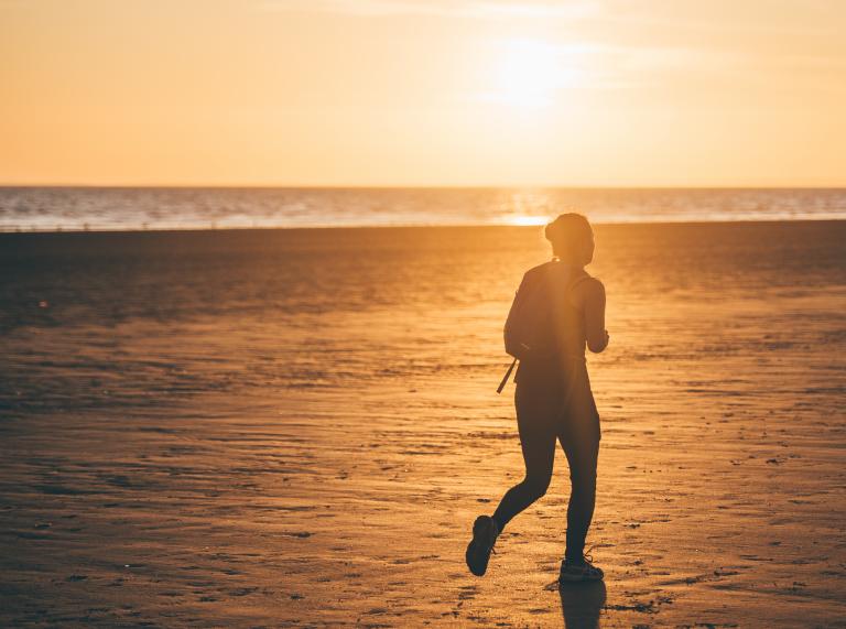 Eine Person, die bei Sonnenuntergang über Sand läuft, mit dem Meer im Hintergrund.