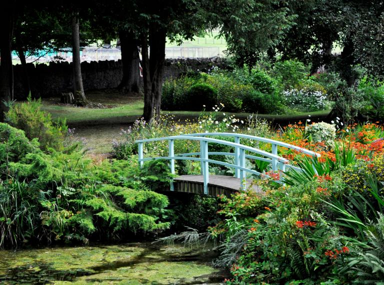 Eine kleine Brücke über einen Teich in einem Garten mit Bäumen und Blumen.