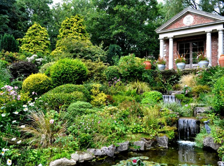Ein üppig grüner Garten mit kleinen Wasserkaskaden und einem Gebäude aus roten Backsteinen und Säulen im Hintergrund.