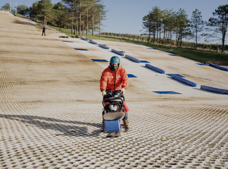 A child in an adapted sled / ski-pram being pushed by a man on a dry ski-slope.