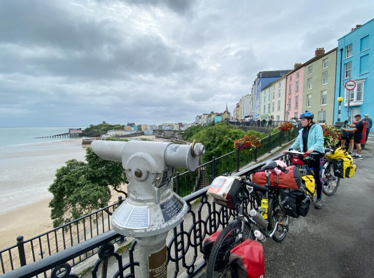 Fahrräder und Menschen stehen entlang des Geländers und blicken auf den Strand und das Meer.