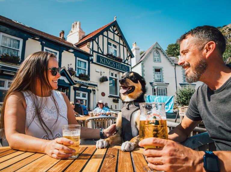 Two people and a dog sitting on a table outside a pub on a sunny day.