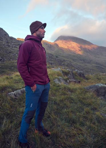 man wearing hat stood in valley with hill in background.