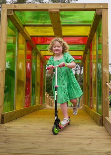 A girl on a scooter in a coloured glass lined wood tunnel.