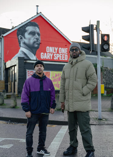 Two men standing in front of a mural of a man on the side of the building. The text reads 'Only one Gary Speed'.