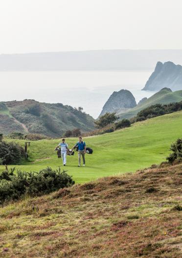 Golfer auf dem Weg zum nächsten Loch auf dem Gelände des Pennard Golf Clubs mit Blick auf die Küste im Hintergrund.