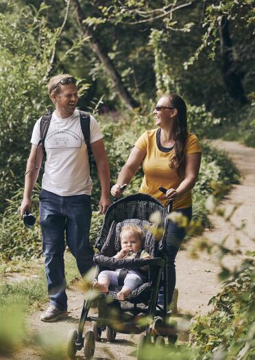 man and woman with baby in pushchair walk along path with greenery and lake in background.