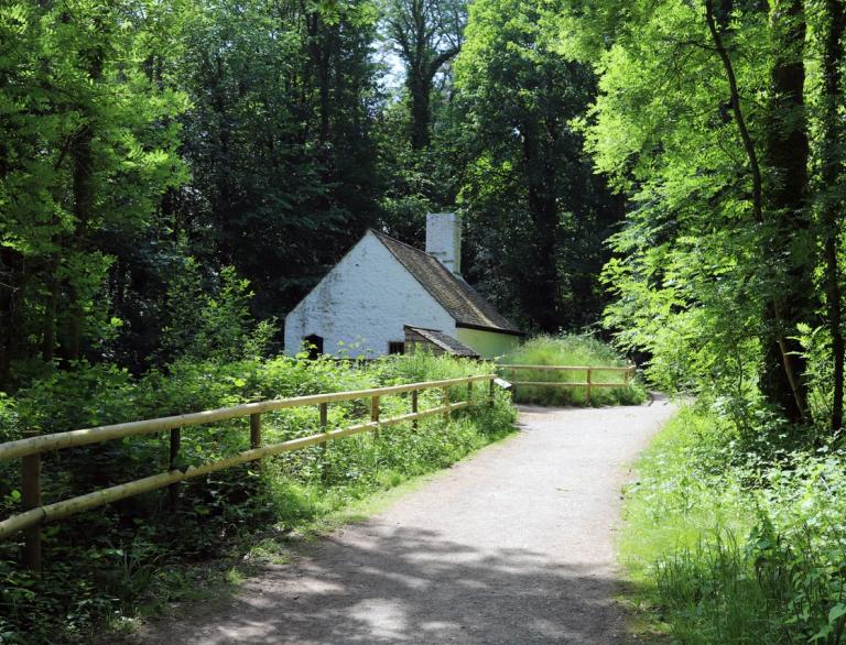 An old white cottage at the end of a path surrounded by greenery.