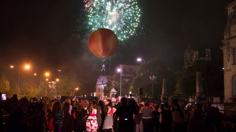 Crowd watch a giant peach in the sky against a back drop of fire works