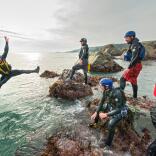 group of male adults laughing as they jump off the rocks into the blue sea, coasteering with instructors.