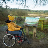 Eine Frau in einem Rollstuhl, die auf eine Informationstafel an einem See blickt.