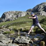 Mann springt über einen Fluss mit Steinen und blauem Himmel im Hintergrund.