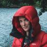 Head and shoulder shot of a lady stood beside a river. She is wearing a red hooded padded jacket with the hood of the coat up,
