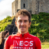 Head and shoulder shot of cyclist Geraint Thomas wearing a red cycling jersey with a grassy bank and part of a stone castle behand him.