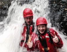 Two adults with red safety helmet and buoyancy aid standing under a waterfall. Female has thumbs up.
