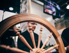 Traditional wooden spinning wheel in the foreground with a person sat behind seen through the spindles of the wheel