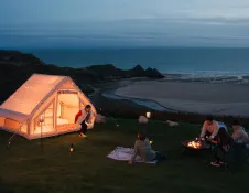 A tent at night time, with a family sat outside round a fire pit, overlooking a beach and bay.