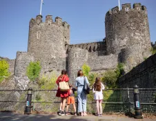 Three visitors standing near a railing and looking up at Conwy Castle’s stone towers under a bright blue sky