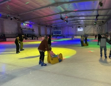 People skating in an indoor ice rink with colourful lights cast across the ice. Some adults are pushing children seated on yellow skating aids, while others skate around the rink.
