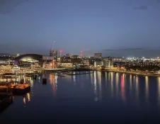 Evening panoramic view of Cardiff Bay with illuminated waterfront buildings and reflections shimmering across the calm water.