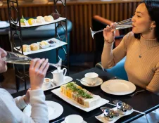 Afternoon tea with sandwiches, scones and cakes on a tiered stand, as two people sit at a table drinking sparkling wine in a modern dining setting.