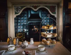 Afternoon tea set with sandwiches, scones and cakes on a three-tier stand beside a lit fireplace, with teacups and silver teapots on the table in a traditional panelled room.