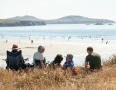People relaxing in coastal grassland overlooking a wide sandy beach and sea, with a headland in the distance