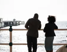 Two people standing by a seaside promenade railing, looking out across a calm bay with a pier and waterfront buildings visible in the background