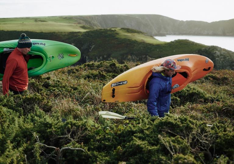 Two people walking through green foliage on a coastal path carrying a kayak each, heading to the sea.