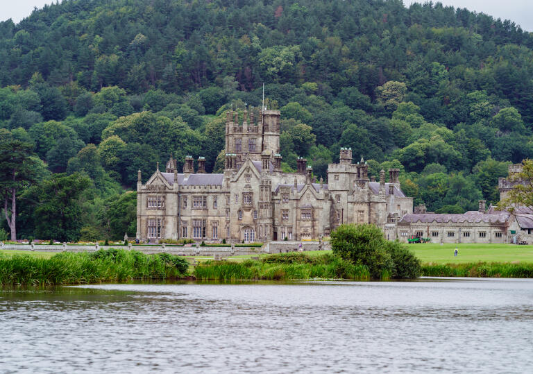 A castle in a parkland with a lake in the foreground.