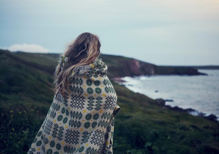 A woman in a blnaket looks out to the coast at sunset