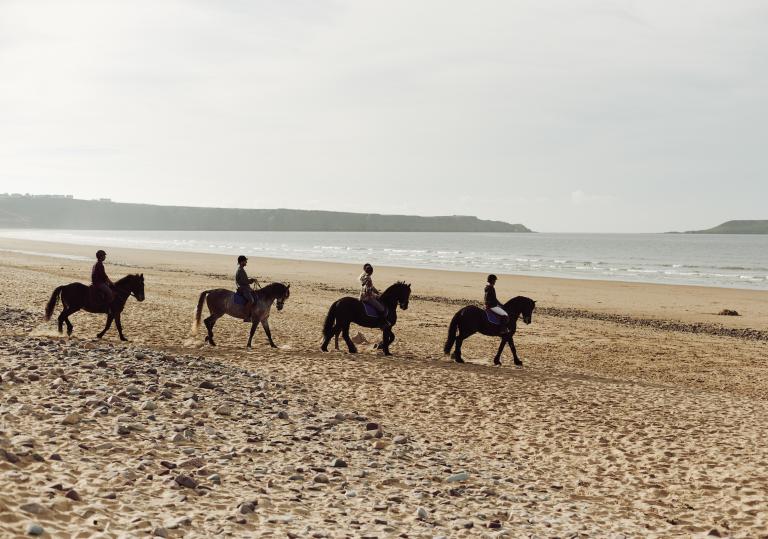 Four horses and riders on a sandy beach.