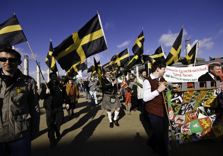 A band marches with flags of St David (which are black with a yellow cross).