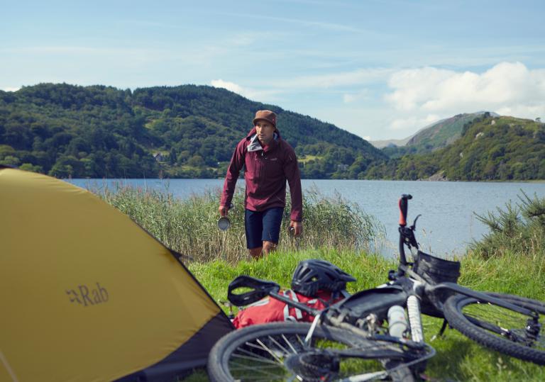 A man in outdoor clothing with a tent and a bike by a lake.