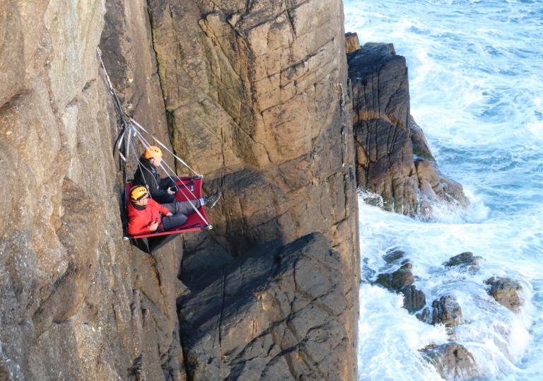 A cliff face above the sea with two people sitting on a portaledge suspended from the cliff edge.