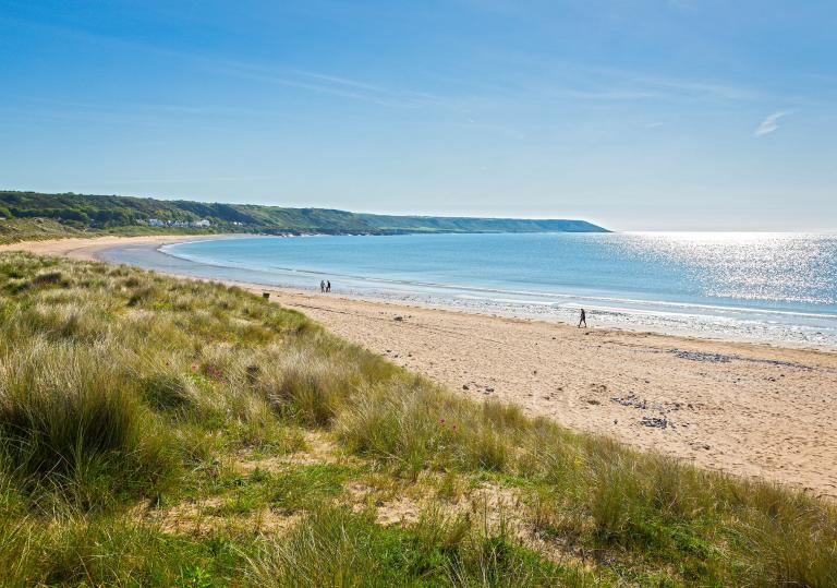 A wide almost deserted beach with grass in the foreground and views of a headland