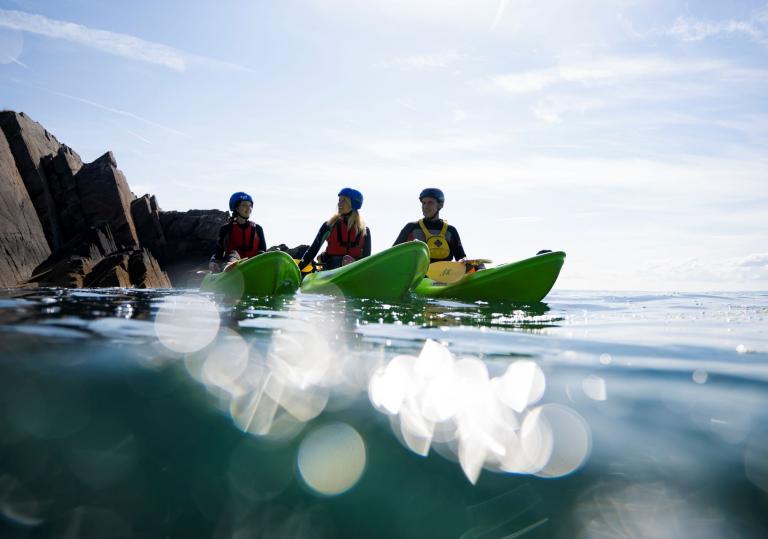 Three kayakers on the ocean with a rocky cove backdrop.