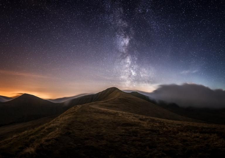 Dark sky full of stars above the mountain landscape of the Brecon Beacons with the orange glow of the setting sun on the horizon.