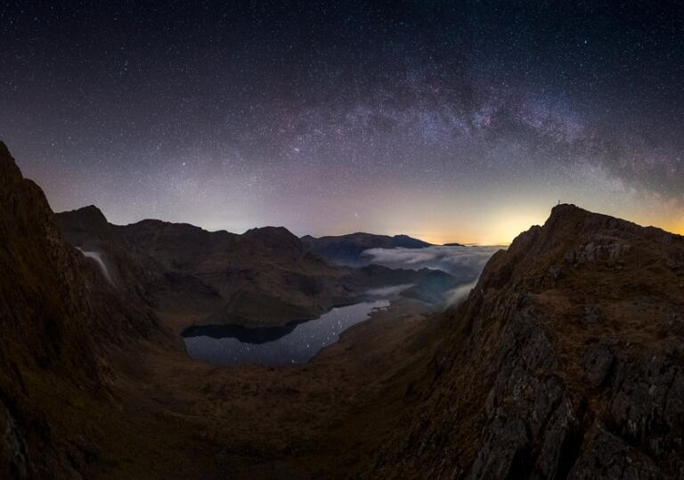 Mountain peaks in Snowdonia silhouetted against a night sky with stars and the orange glow of sunset on the horizon.