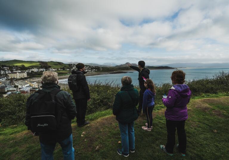 A tour guide with a group, pointing to a seaside town and castle from a cliff top.