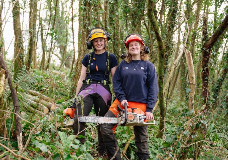 Two women in protective clothing holding chainsaws in a forest.